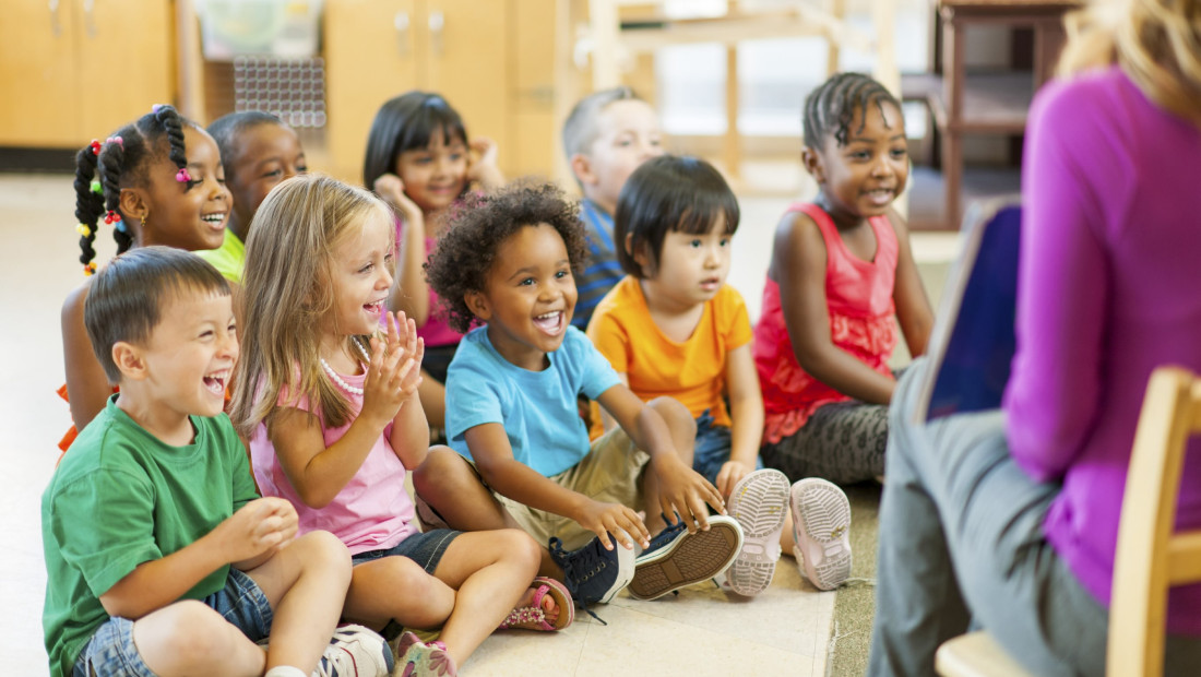 Group of kindergarten children sitting in front of a teacher reading a picture book to the class