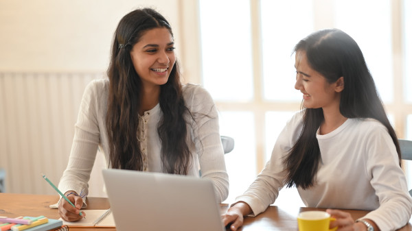 Student tutoring another student at a desk with notes and a laptop