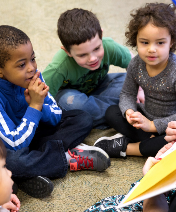 children sitting around teacher who is reading a picture book aloud